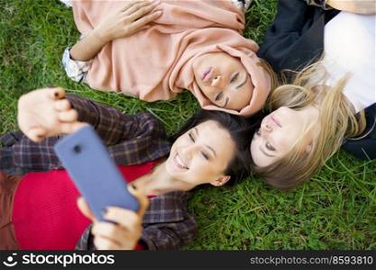Top view of multiethnic young ladies lying on grass and taking self portrait on cellphone. Multiracial girlfriends lying in circle and taking selfie