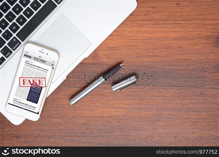 Top view of office desk table with laptop computer, pen and smartphone phone with digital fake News on wooden background. Digital newspaper concept. All screen graphics are made up by us