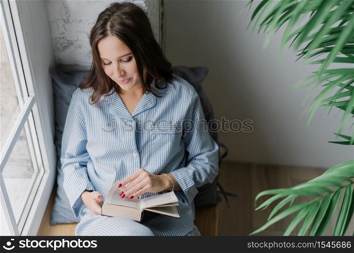 Top view of pleased woman with dark long hair, focused into book, reads something exciting, enjoys cozy atmosphere at home, poses on window sill in empty room. Its time for reading and rest.