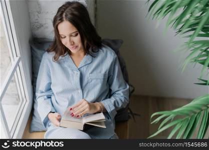 Top view of pleased woman with dark long hair, focused into book, reads something exciting, enjoys cozy atmosphere at home, poses on window sill in empty room. Its time for reading and rest.