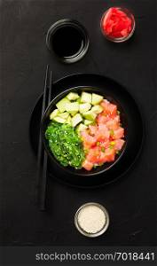 Top view of poke with diced red fish, avocado and green seaweeds salad decorated with green onions and sesame seeds in black bowl with soy sauce and ginger on dark background.. Top view of poke with diced red fish, avocado and green seaweeds salad.