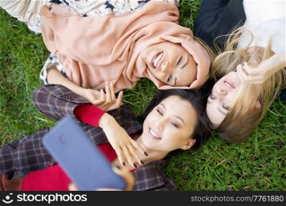 Top view of positive diverse female friends taking self portrait while lying together on green lawn on summer day in park. Multiracial women taking selfie together in park