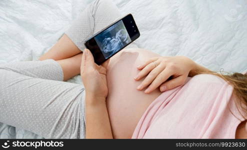 Top view of pregnant woman holding smartphone and looking on ultrasound image of her unborn baby in belly. Concept of expecting baby, pregnancy and healthcare. Top view of pregnant woman holding smartphone and looking on ultrasound image of her unborn baby in belly. Concept of expecting baby, pregnancy and healthcare.