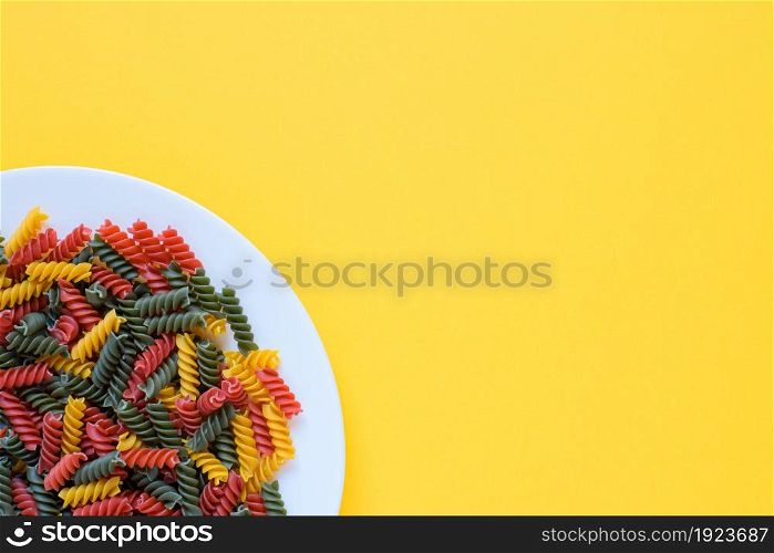 Top view of red green and yellow dry raw fusilli pasta on a plate with yellow background and copy space.. Red green and yellow dry raw fusilli pasta on a plate with yellow background and copy space.