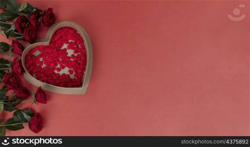 Top view of red rose flowers and a heart shaped gift box filled with tiny hearts forming left border on a soft red background for a Valentines day holiday concept