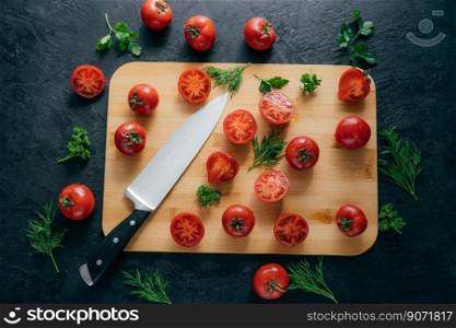 Top view of red sliced tomatoes on wooden chopping board. Sharp knife near. Green parsley and dill. Dark background. Preparing fresh vegetable salad