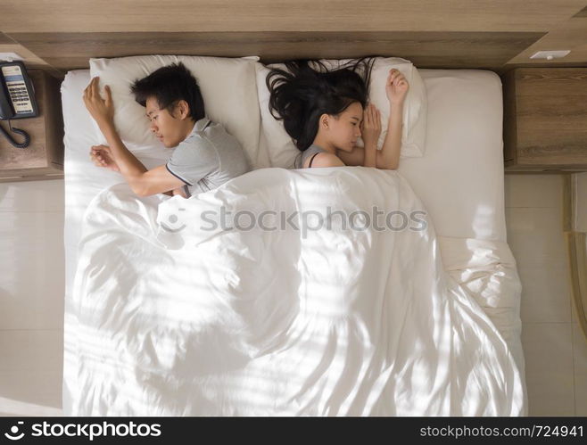 Top view of sad Asian couple sleeping together, thinking about relationship problems, and suffering from depression on bed with white blanket,