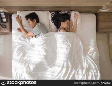 Top view of sad Asian couple sleeping together, thinking about relationship problems, and suffering from depression on bed with white blanket,