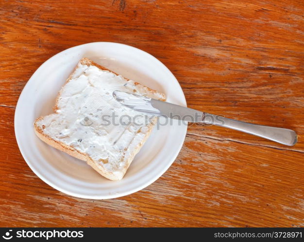 top view of sandwich from toast and soft cheese with herbs on white plate, table knife on wooden table