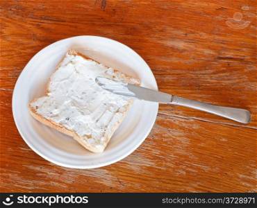 top view of sandwich from toast and soft cheese with herbs on white plate, table knife on wooden table