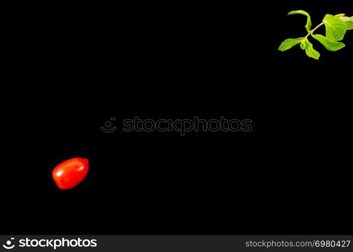 Top view of single cherry tomato on one side and mint leaves diagonally opposite isolated on a black background. Minimalist picture style with copy space for text