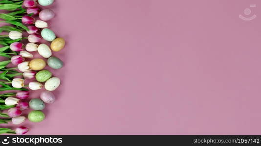 Top view of springtime tulip flowers and egg decorations forming left border on a soft pink background for a happy Easter holiday concept