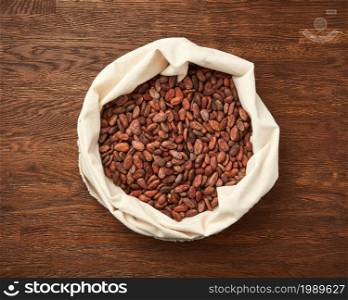 Top view of textile white bag filled with raw unpeeled cocoa beans placed on rustic wooden surface. Sack of organic unpeeled cocoa beans