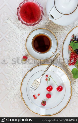 Top view of the beautifully decorated table with cup of tea, white plates with different berries, linen napkin, cutlery and jam on luxurious tablecloths