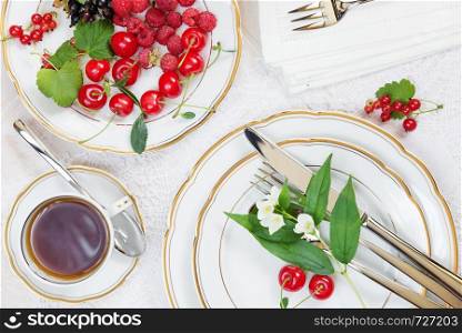 Top view of the beautifully decorated table with cup of tea, white plates with different berries, linen napkin, cutlery and flowers on luxurious tablecloths
