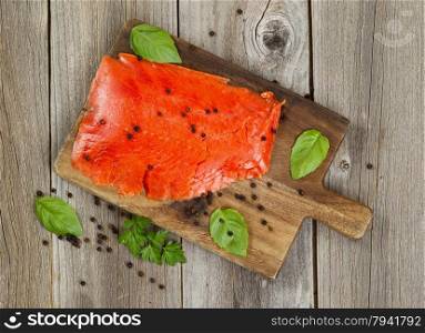 Top view of thinly sliced cold smoked red salmon along with herbs and seasoning on wooden server board. Rustic wood underneath.