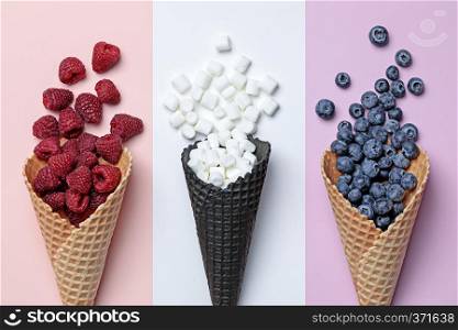 Top view of three waffle cones with ripe berries and marshmallows on pastel color block backgrounds, flat lay