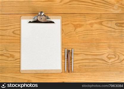 Top view of traditional clipboard, blank paper, and pens on wooden desktop.