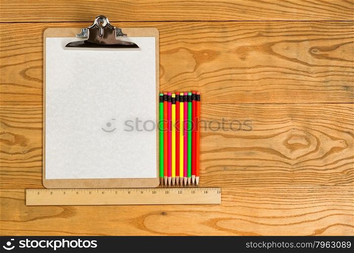 Top view of traditional clipboard, blank paper, ruler, and colorful pencils on wooden desktop.