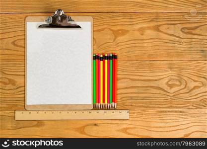 Top view of traditional clipboard, blank paper, ruler, and colorful pencils on wooden desktop.