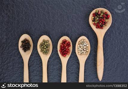 Top view of various colors peppercorns in wooden spoon on black stone slate background