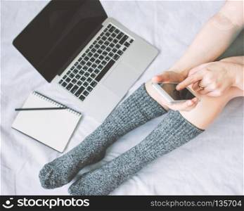 Top view of woman legs in socks and using smartphone and laptop on cozy bed, lifestyle concept, autumn and winter season