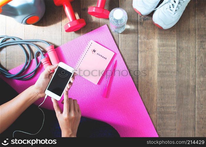 Top view of woman using mobile phone for online fitness training with fitness equipments on wooden floor
