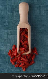 Top view of wooden scoop with dry red goji berries against blue vinyl background.