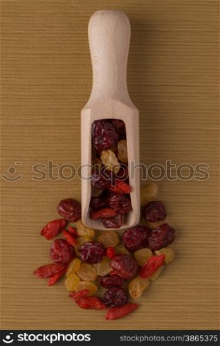 Top view of wooden scoop with mixed dried fruits against brown vinyl background.