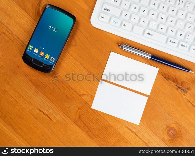 Top view on wooden table with white keyboard, smart phone with green screen and business cards. Desk with keyboard, smartphone and business card