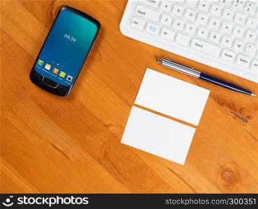 Top view on wooden table with white keyboard, smart phone with green screen and business cards. Desk with keyboard, smartphone and business card
