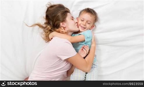 Top view portrait of little boy in pajamas hugging and embracing his smiling mother lying with him on bed with white sheets. Concept of parenting, loving children and family happiness.. Top view portrait of little boy in pajamas hugging and embracing his smiling mother lying with him on bed with white sheets. Concept of parenting, loving children and family happiness