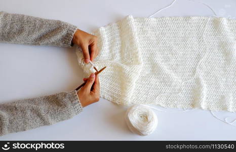 Top view simple concept for wintertime, woman hand with knitting needles to knit white scarf for handmade seasonal gift on white background, amazing gentles color