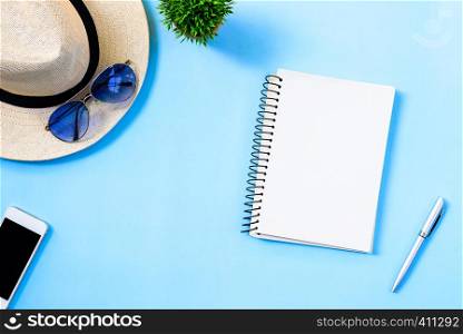 Top view summer travel planning concept with white hat, blue eyeglasses, phone and notebook isolated on blue background.