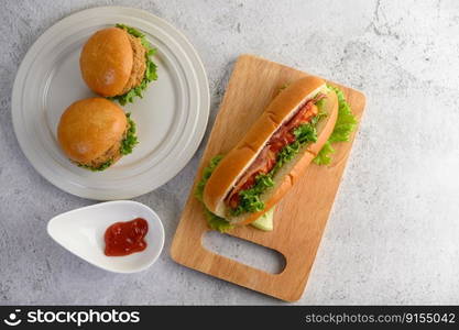 Top view with two hamburgers in white dish and large hotdog on wooden cutting board, tomato sauce and lettuce on floor, copy space