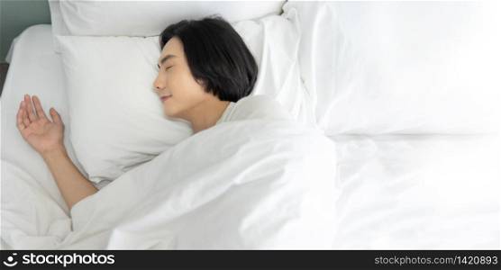 Top view Young man sleeping and dreaming in white bed in the morning. Happy smiling Asian male enjoying lying on comfortable soft bedding in bedroom at home. eyes closed. copy space.