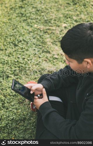 top view young millennial boy sitting in the park, in green Scutch grass with black casual clothes, looking at his smart phone, selective focus, concept of technology.. top view young boy sitting in the park, looking at his smart phone, selective focus