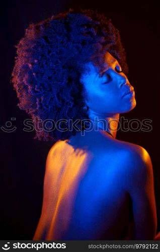 Topless black woman with Afro hairstyle closing eyes under orange and blue neon light against black background. Naked African American model under neon illumination