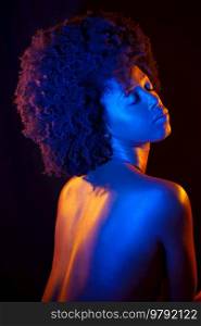 Topless black woman with Afro hairstyle closing eyes under orange and blue neon light against black background. Naked African American model under neon illumination