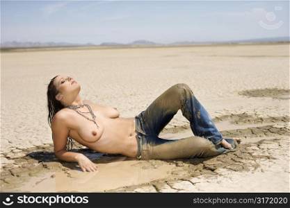 Topless Caucasian mid-adult woman covered in mud lying in desert with head back.