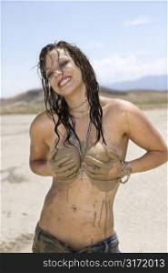 Topless Caucasian mid-adult woman holding breasts covered in mud in desert smiling and looking at viewer.