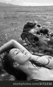 Topless Caucasian mid adult woman on rocky Maui coast lying on back with eyes closed.