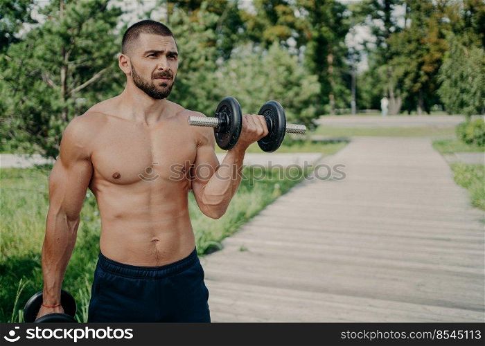 Topless weightlifter raises heavy barbell enjoys strength training, has healthy lifestyle, focused into distance, exercises outdoor, lifts up dumbbell, being in good physical shape. Sport concept