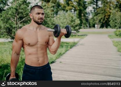 Topless weightlifter raises heavy barbell enjoys strength training, has healthy lifestyle, focused into distance, exercises outdoor, lifts up dumbbell, being in good physical shape. Sport concept