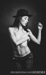 Topless woman wearing blue jeans, panties and hat on black background. Black and white studio photograph.