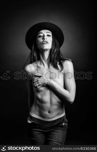 Topless woman wearing blue jeans, panties and hat on black background. Black and white studio photograph.