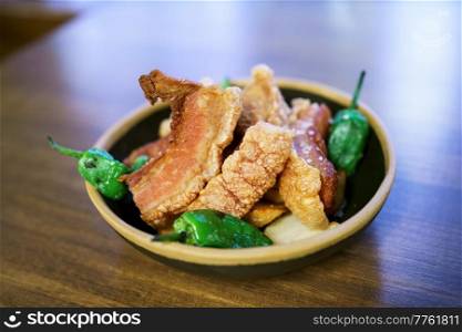 Torreznos, pieces of deep fried, meat with green hot peppers served in bowl on lumber table in restaurant. Fried meat served in bowl with peppers