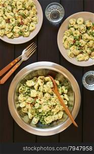 Tortellini salad with green peas, fried bacon and parsley in big salad bowl with spoon to serve, salad served on two plates, forks and glasses, photographed overhead on dark wood with natural light