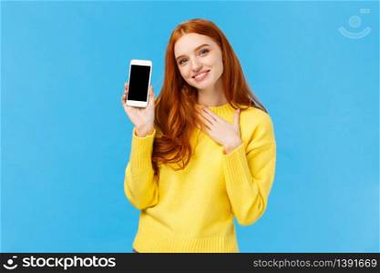Touched lovely and romantic redhead cute woman bragging with photos from winter holidays, showing smartphone display, touch heart and sighing grateful or pleased, blue background. Copy space. Touched lovely and romantic redhead cute woman bragging with photos from winter holidays, showing smartphone display, touch heart and sighing grateful or pleased, blue background