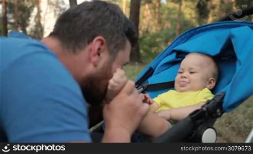 Touching portrait of affectionate stylish dad with infant son in pram kissing his feet outdoors. Caring father kissing toddler baby boy&acute;s feet while enjoying time together in summer park. Side view. Closeup. Slow motion. Stabilized shot.